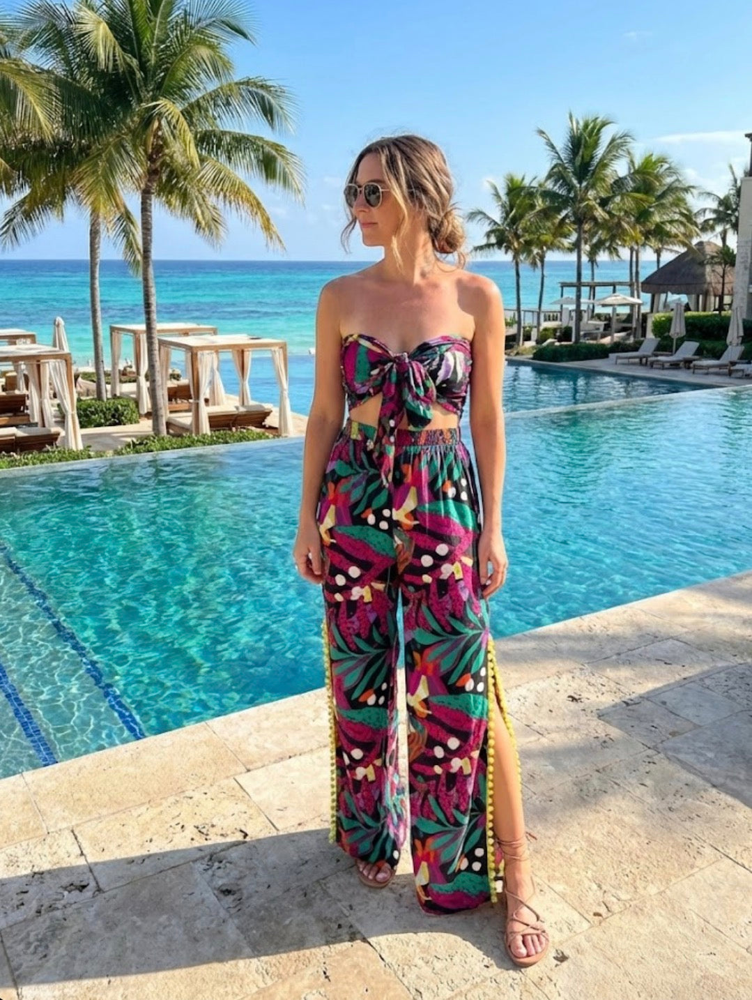 Woman in a colorful matching crop top and pants standing by a pool with palm trees and ocean in the background