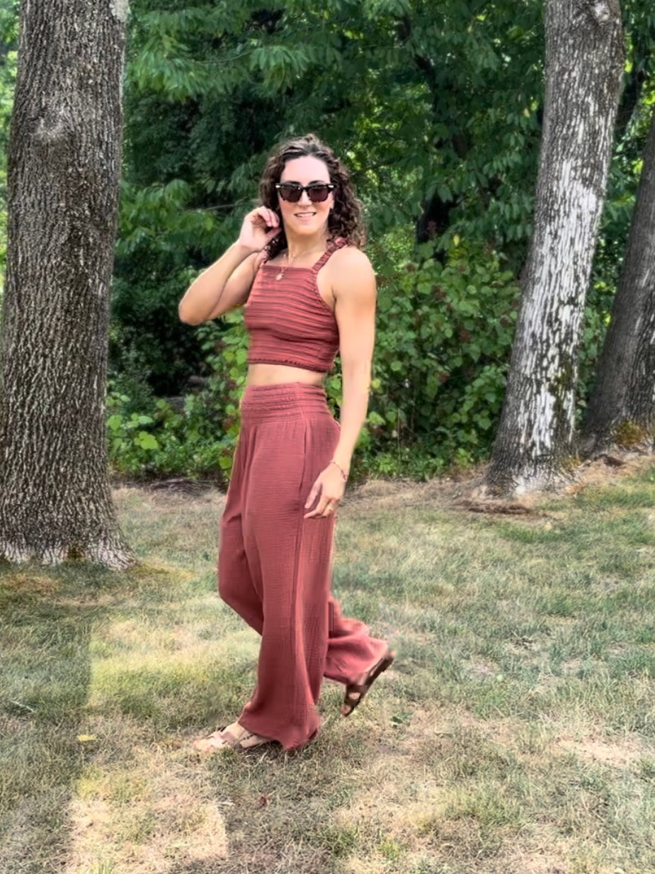 A woman in a rust gauzy palazzo outfit is standing in a park with trees and grass.