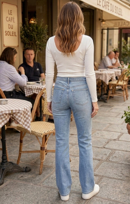 Woman in light blue jeans and white shirt walking on a street with people sitting at tables in the background.