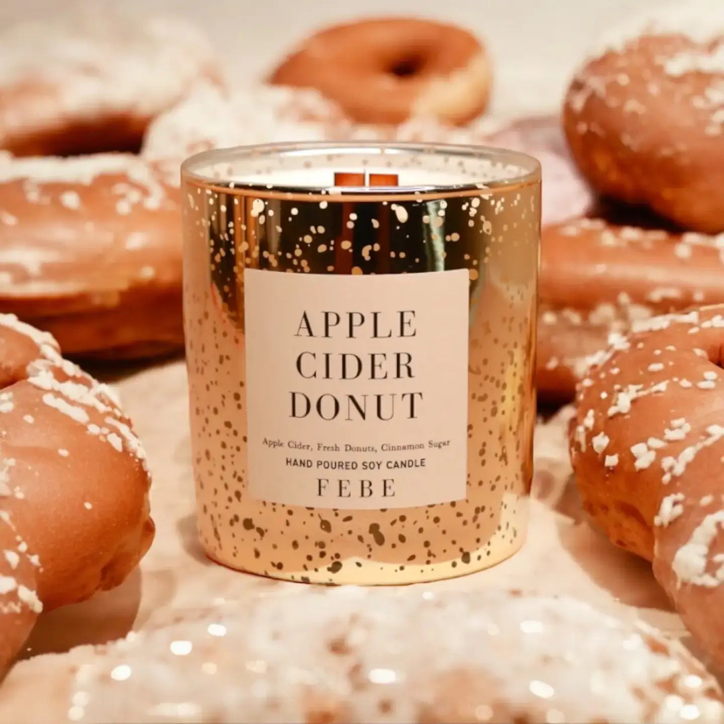 Candle in a gold container labeled 'Apple Cider Donut' by FEBE surrounded by donuts