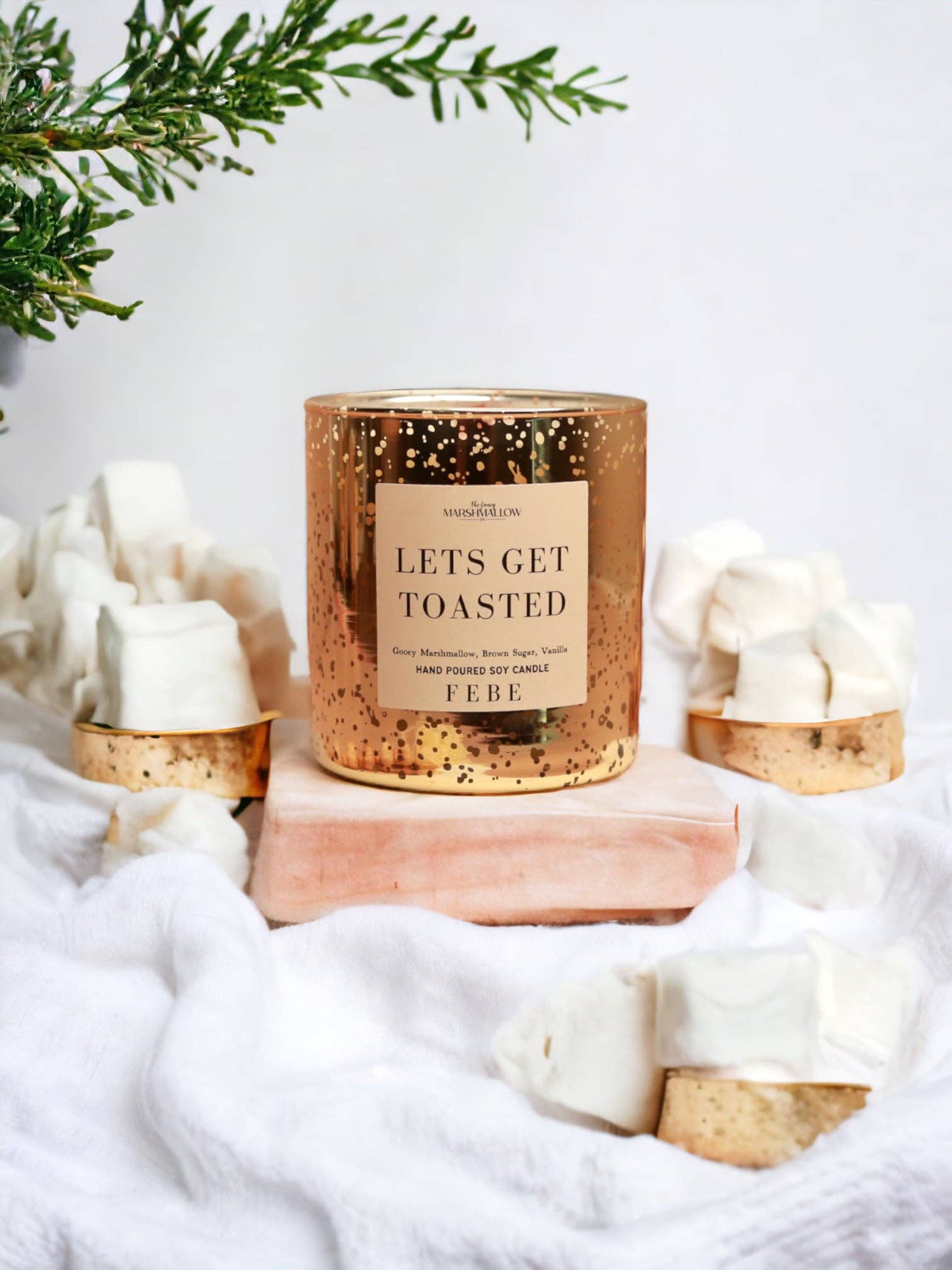 Candle labeled 'Let's Get Toasted' by FEBE with marshmallows and Christmas decorations on a white background
