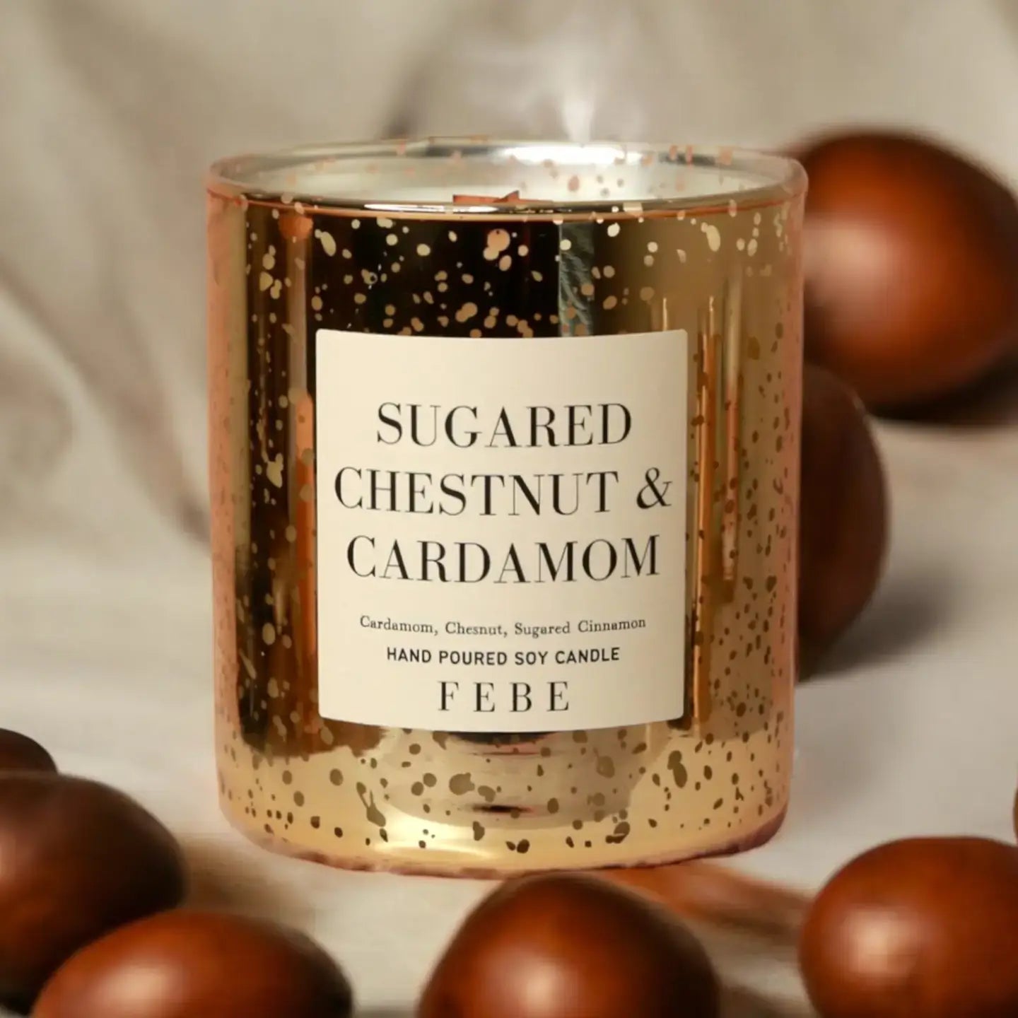 Candle in gold container with 'Sugared Chestnut & Cardamom' label surrounded by chestnuts