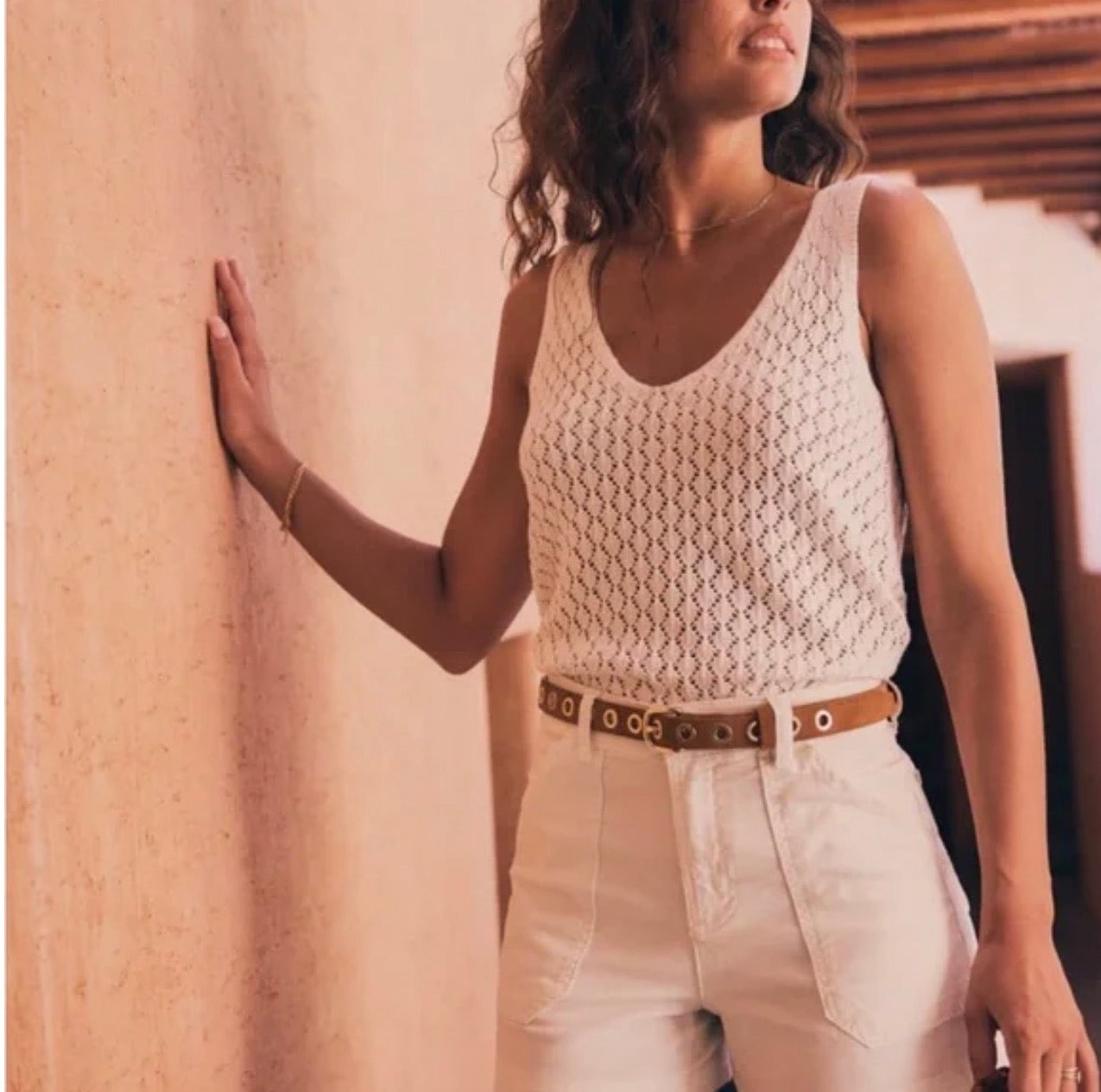 Woman wearing a white crochet top and light-colored pants against a beige wall.