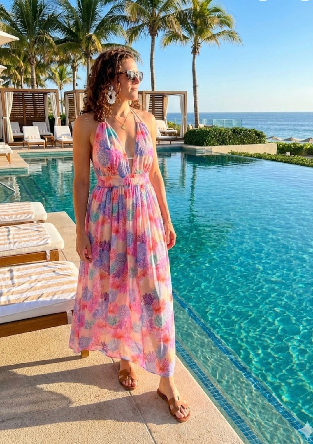Woman in a colorful dress standing by a pool with palm trees and ocean in the background