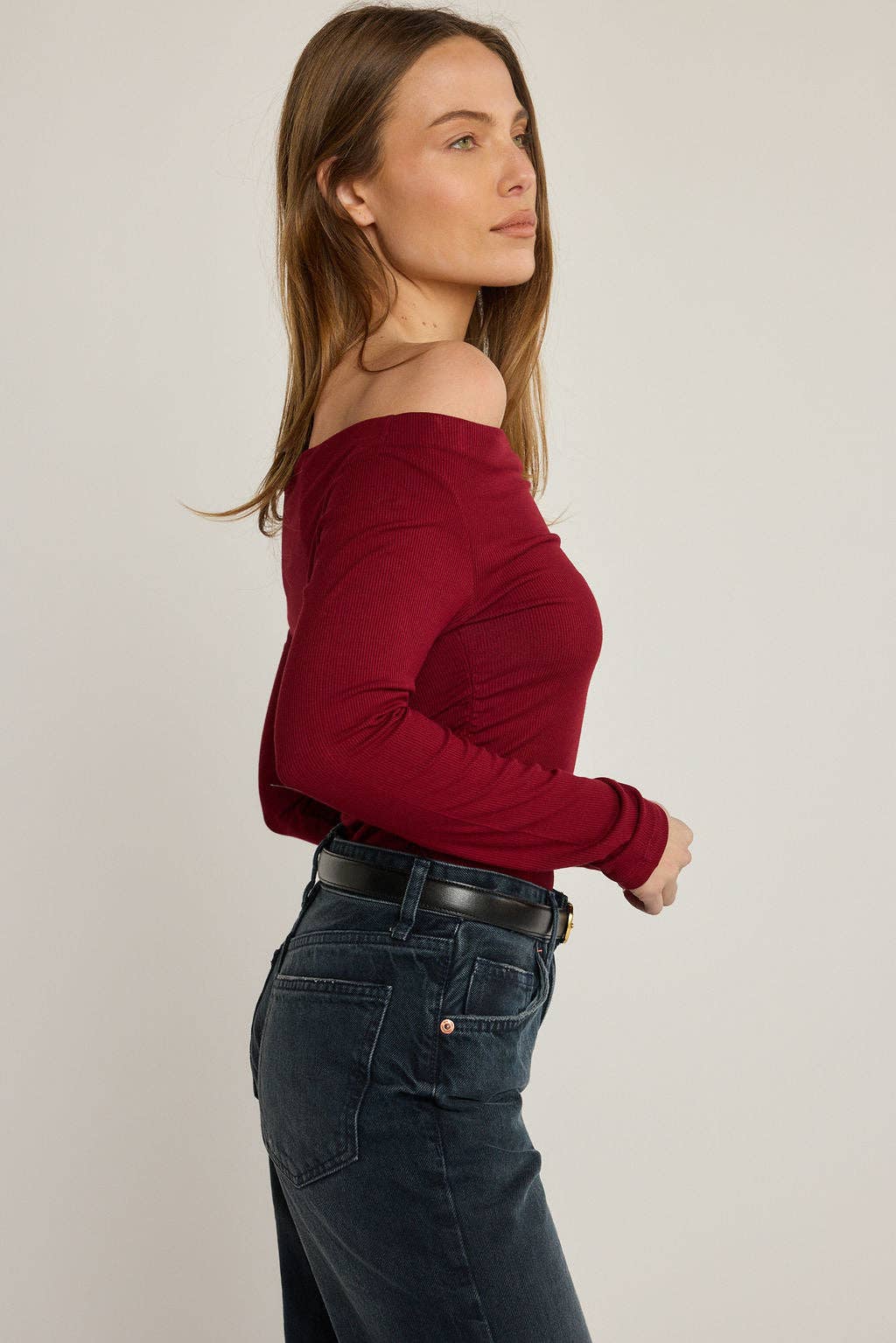 Woman wearing a red off-shoulder top and dark jeans against a plain background
