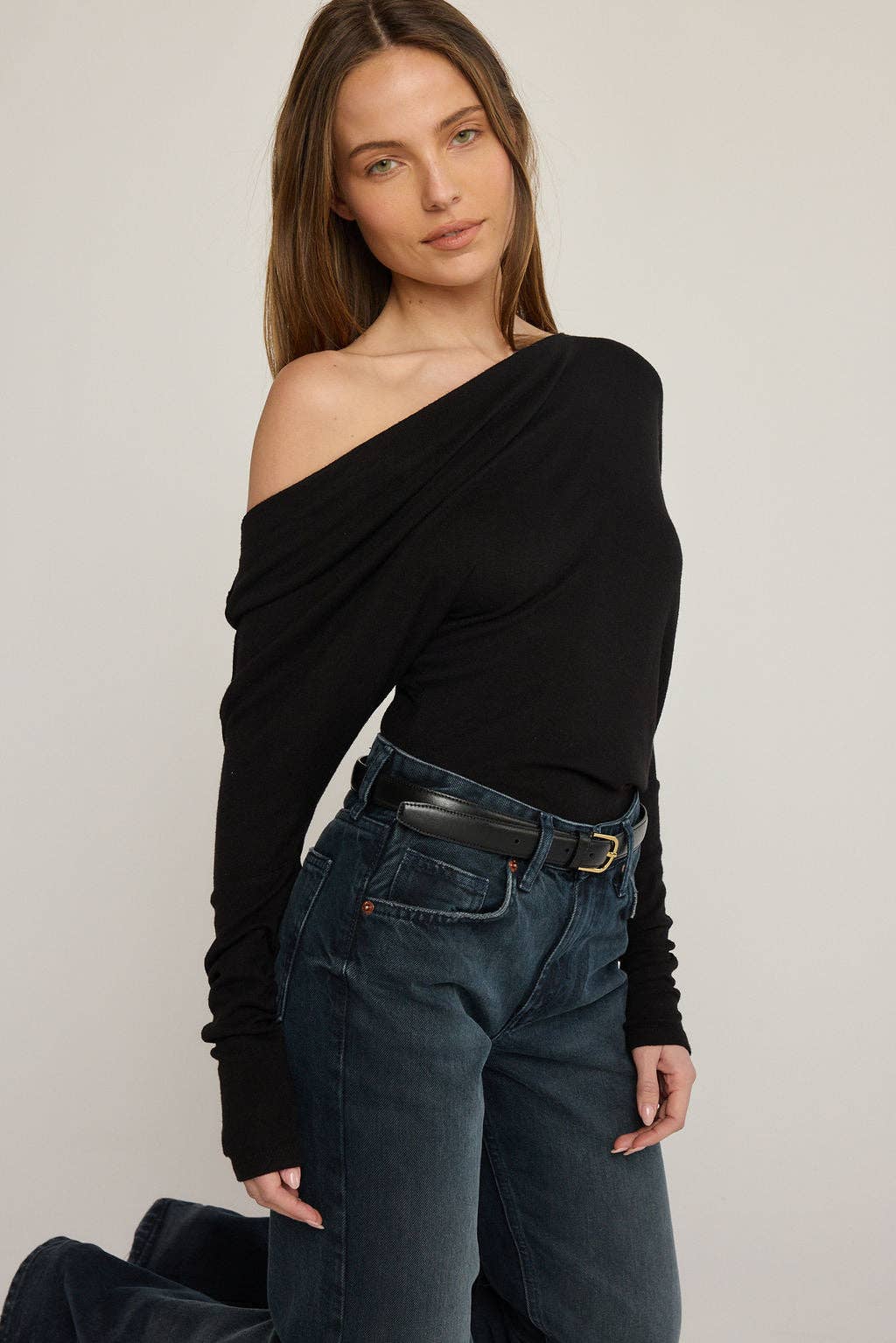Woman wearing a black off-shoulder top and dark jeans against a plain background