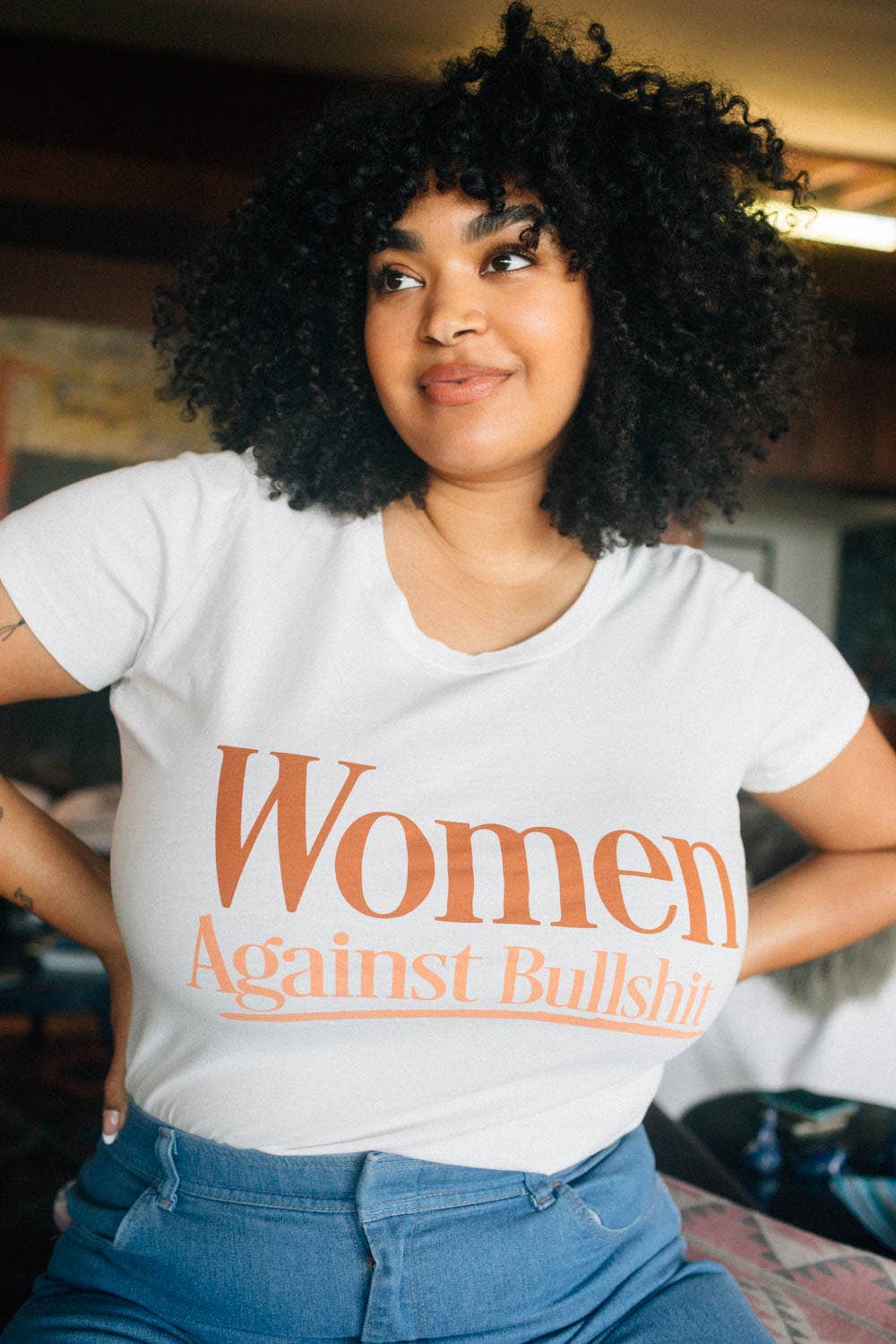 A woman wearing a white scoop neck t-shirt with the text 'WOMEN AGAINST BULLSHIT' printed on the front.